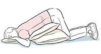Hombre acostado boca abajo con almohadas debajo de la cabeza y del abdomen.